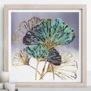 Op de afbeelding: Een ingelijst diamanten schilderij van een ginkgo-blad in tinten groen, blauw en goud. De bladeren zijn in een waaiervorm gerangschikt en zijn geplaatst op een grijze achtergrond.