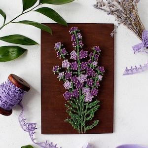 Op de afbeelding: Een houten plaquette met een string art ontwerp van paarse en groene lavendelbloemen. De plaquette is omringd door lavendeltakken, kant, een spoel paarse kant, groene bladeren, een gieter en een paarse gieter.