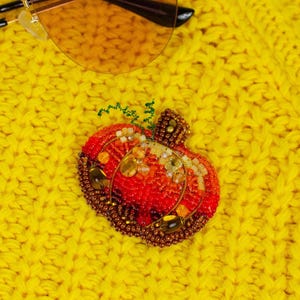 May include: A pumpkin-shaped brooch crafted from intricate beadwork in shades of red, orange, and brown. The design features a textured surface with metallic accents, clear crystal embellishments, and a small green wire stem detail on top.