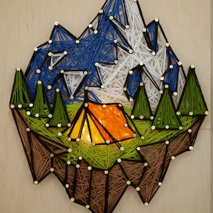 May include: A string art piece depicting a mountain scene with a tent. The mountains are blue and white, the trees are green, and the tent is orange. The artwork is mounted on a wooden board.