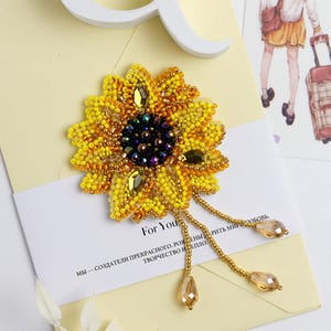 Könnte beinhalten: Eine bestickte Sonnenblumenbrosche mit gelben, goldenen und dunkelblauen Perlen. Die Brosche hat goldene Ketten mit tropfenförmigen Perlen. Die Brosche liegt auf einer weißen Karte mit dem Text "For You". Die Karte liegt auf einem gelben Umschlag.