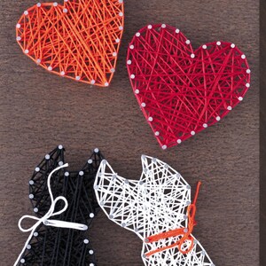 Puede incluir: Arte de hilo con dos corazones y dos siluetas de gatos. Los corazones están hechos con hilo naranja y rojo, mientras que los gatos son negros y blancos. La obra de arte está montada sobre una superficie de madera marrón.