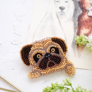May include: A handcrafted beaded pug brooch. The pug is depicted in shades of brown, tan, and black beads, with large, expressive eyes. The brooch is set against a white background with a watercolor illustration of dogs in the background.