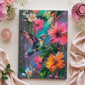 May include: A diamond painting kit featuring a vibrant hummingbird in flight surrounded by large pink and orange hibiscus flowers and smaller white blossoms against a dark grey background, created with colourful faceted resin beads.