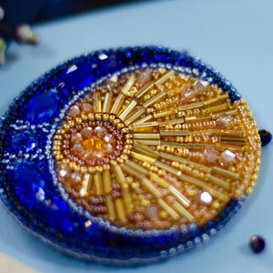 Könnte beinhalten: Handgefertigte Brosche mit einem Himmelsdesign. Die Brosche hat eine halbmondförmige Form in tiefem Blau, die ein Sonnenstrahl-Design aus goldenen und orangefarbenen Perlen umschließt. Die Brosche ist mit glitzernden Kristallen und Perlen verziert.