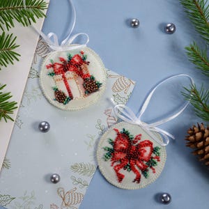 May include: Two white round ornaments with red and green beaded designs. One ornament features a gift box with a red bow and the other features a red bow with green leaves.