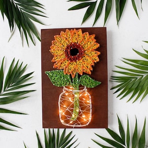 May include: A handmade string art piece featuring a sunflower in a mason jar. The sunflower has orange and yellow petals with a dark center. The jar and flower stem are green, and the jar is illuminated with fairy lights, all on a brown wooden background.