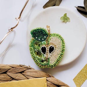 May include: A handmade, beaded apple brooch, cut in half, with detailed beadwork in green and cream. The brooch has a leaf and black seed details, presented on a white plate. A unique, handcrafted accessory.