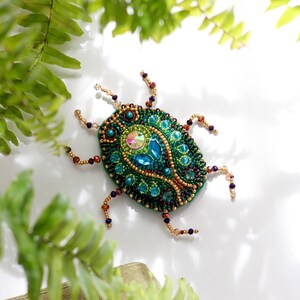 May include: A handcrafted, beaded insect brooch in shades of teal, green, and gold. The bug-shaped pin features intricate beadwork, including a central iridescent gem and blue teardrop-shaped stones. The brooch is set against a white background with green foliage.