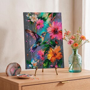 May include: A diamond painting canvas featuring a hummingbird in flight surrounded by vibrant pink and orange hibiscus flowers against a dark grey background, displayed on a gold metal easel stand with a small dish of colourful beads.