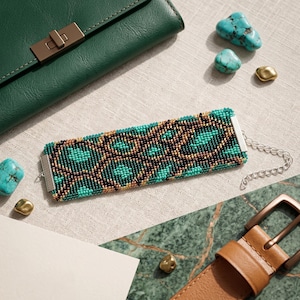 Può includere: Un bracciale rettangolare di perline con un motivo geometrico nei toni del turchese, verde scuro e bronzo metallizzato. Il bracciale presenta terminali in metallo argentato e una catenella di estensione argentata con chiusura a moschettone.