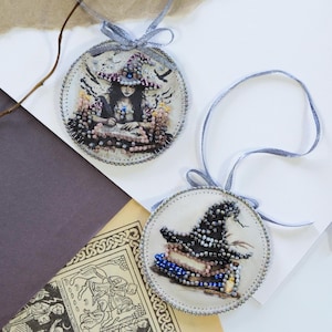 Può includere: Due ornamenti circolari da appendere con intricate decorazioni di perline. Uno raffigura una strega che scrive su un libro, l'altro un cappello da strega su una pila di libri. Entrambi sono rifiniti con nastri argentati per essere appesi.