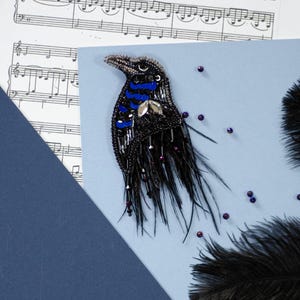 May include: A handcrafted raven brooch with a beaded design. The bird features black beads, blue accents, and a silver-coloured gem. Black feathers and small purple beads add detail. The brooch is set against a light blue background with sheet music.