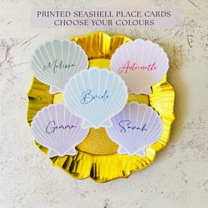 Peut inclure: Cartes de table en forme de coquillage, imprimées en couleurs pastel, disposées sur une assiette dorée. Chaque carte porte un nom écrit en script. Le texte en haut indique : "PRINTED SEASHELL PLACE CARDS CHOOSE YOUR COLOURS."