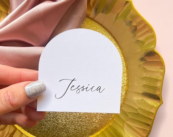 May include: White place card with a rounded top and the name "Jessica" written in cursive script. The place card is sitting on a gold charger plate.