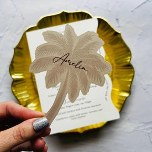 May include: A beige palm tree-shaped place card with the name "Amelia" in cursive script. The place card is set against a gold-coloured, scalloped-edge plate, with a white menu card visible behind it.