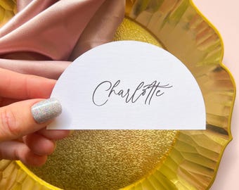 May include: White half-circle place card with the name "Charlotte" written in cursive on a gold charger.