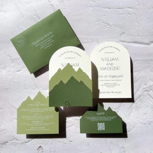 May include: A collection of olive green wedding stationery, including an envelope, invitations, and RSVP cards. The invitations feature a mountain design and the names "William and Madeline". The envelope reads "Please Deliver To".