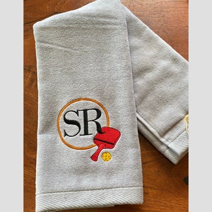 May include: A grey towel with a personalised embroidery design featuring the initials "SR" in a circle with a red and yellow paddle and ball.