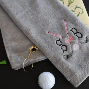 May include: A gray golf towel with embroidered golf club and glasses design, a pink bow tie, and the initials "SB". Includes golf balls, green tees, and a gold towel ring.