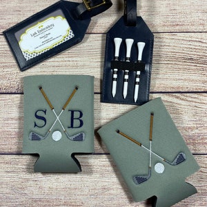 Op de afbeelding: Twee groene en grijze koelers voor blikjes met een golfclub- en balontwerp. Een koeler heeft de initialen "SB" geborduurd. Een zwart leren bagagelabel met vier golftees. Een zwart leren bagagelabel met de tekst "Lark Embroidery" en "Stacey Boyle" erop.