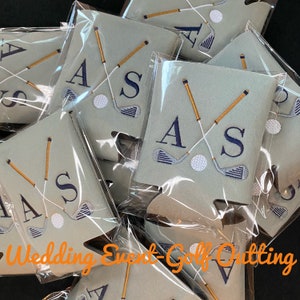 Op de afbeelding: Een stapel gepersonaliseerde golf-thema can coolers in grijs en wit met de initialen "A.S." en een golfclub ontwerp. De can coolers zijn verpakt in doorzichtige plastic zakken. "Wedding Event - Golf Outting" staat in oranje tekst onderaan de afbeelding.