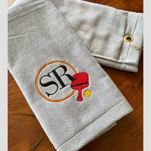 May include: Grey towel with a loop for hanging. The towel has an embroidered design of a circle with the letters "SR" inside, a red paddle, and a yellow ball.