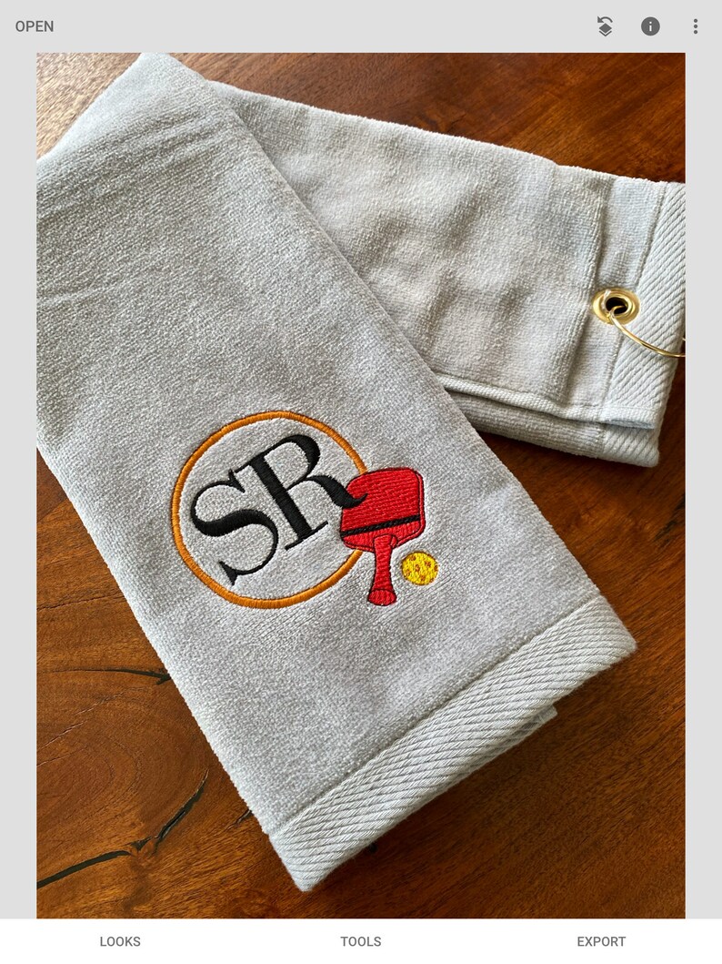 May include: Grey towel with a loop for hanging. The towel has an embroidered design of a circle with the letters "SR" inside, a red paddle, and a yellow ball.