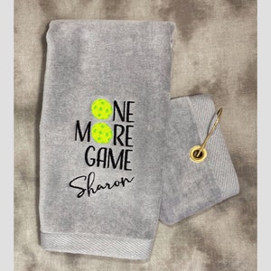 以下が含まれることがあります： グレーのタオルに、「One More Game」という文字と「Sharon」という名前が黒で刺繍されています。テキストの上に2つの黄色のピクルボールのアイコンがあります。タオルには吊り下げ用のループが付いています。