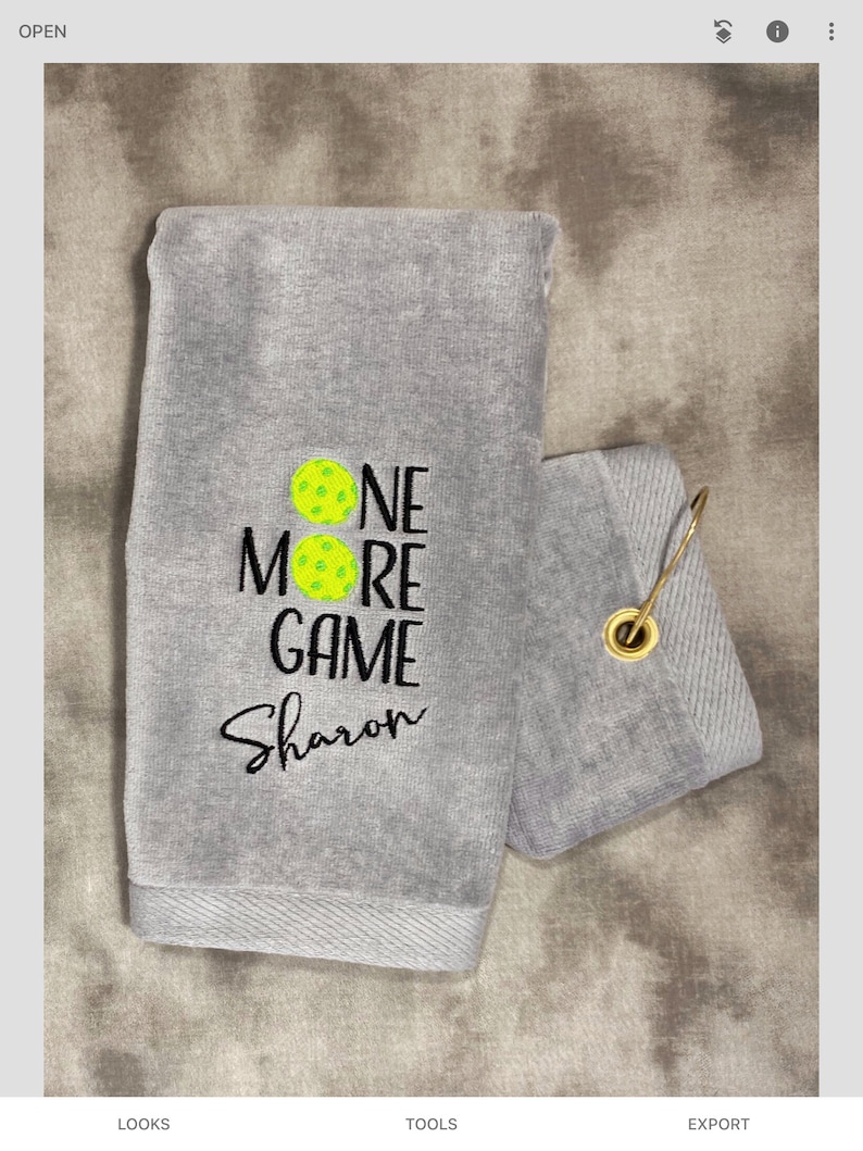 以下が含まれることがあります： グレーのタオルに、「One More Game」という文字と「Sharon」という名前が黒で刺繍されています。テキストの上に2つの黄色のピクルボールのアイコンがあります。タオルには吊り下げ用のループが付いています。