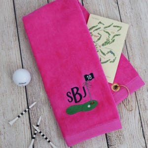 May include: A vibrant pink golf towel with embroidered initials and a golf course design. The towel features a golf flag with the number 18, a golf ball, and golf tees. A golf course map is tucked inside.