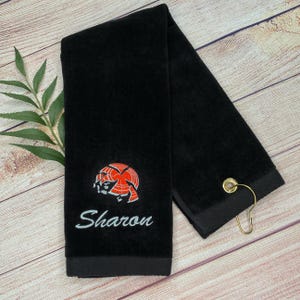 May include: Black golf towel with a red and white clay pigeon design and the name "Sharon" embroidered in white.