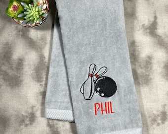 May include: Gray hand towel with a white border. The towel has an embroidered design of two bowling pins and a bowling ball. The name "PHIL" is embroidered in red below the bowling pins and ball.