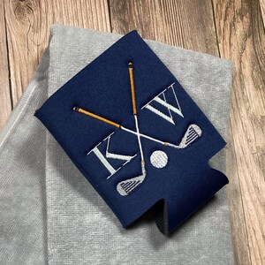 May include: A navy blue can cooler with a white embroidered design of crossed golf clubs, a golf ball, and the initials "KW".