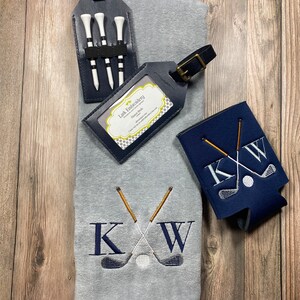 May include: A grey golf towel with the initials KW embroidered in white with crossed golf clubs and a golf ball. A blue and white golf can cooler with the initials KW embroidered in white with crossed golf clubs and a golf ball. A black leather luggage tag with a business card for Lark Embroidery. A black leather golf tee holder with three white golf tees.