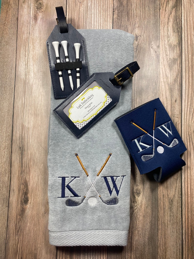 May include: A grey golf towel with the initials KW embroidered in white with crossed golf clubs and a golf ball. A blue and white golf can cooler with the initials KW embroidered in white with crossed golf clubs and a golf ball. A black leather luggage tag with a business card for Lark Embroidery. A black leather golf tee holder with three white golf tees.