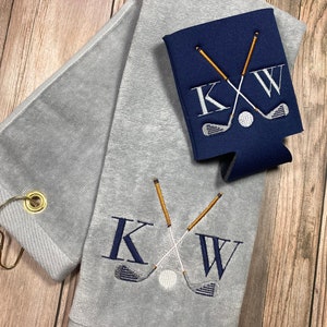 May include: Grey golf towel with a white and gold embroidered design of crossed golf clubs and the initials "KW". A matching blue cooler sleeve with the same design is also shown.