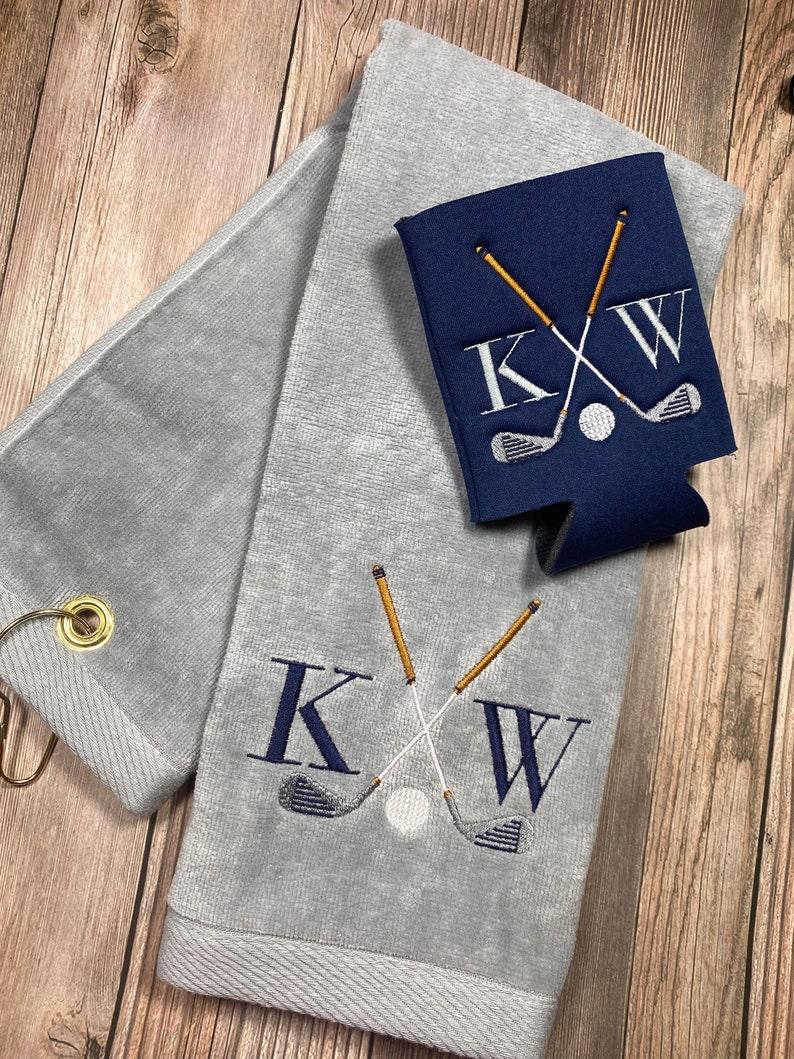 May include: Grey golf towel with a white and gold embroidered design of crossed golf clubs and the initials "KW". A matching blue cooler sleeve with the same design is also shown.