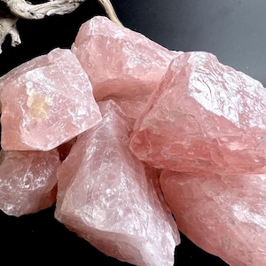 Può includere: Un gruppo di cristalli di quarzo rosa grezzi. Le pietre, di varie dimensioni e forme, presentano una tonalità rosa tenue. I cristalli hanno un aspetto naturale, non lucidato, con texture e sfaccettature visibili. La disposizione suggerisce pietre preziose naturali.
