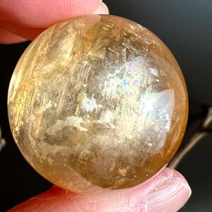 May include: A translucent, golden-yellow sphere with internal fractures and iridescent flashes. The polished crystal ball is held between fingers, showcasing its smooth, rounded form and natural variations. The sphere's surface reflects light, creating a play of colors.