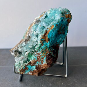Puede incluir: Un esp&eacute;cimen de cristal azul verdoso y marr&oacute;n con formaciones de cristales blancos. El cristal est&aacute; en un soporte de metal.