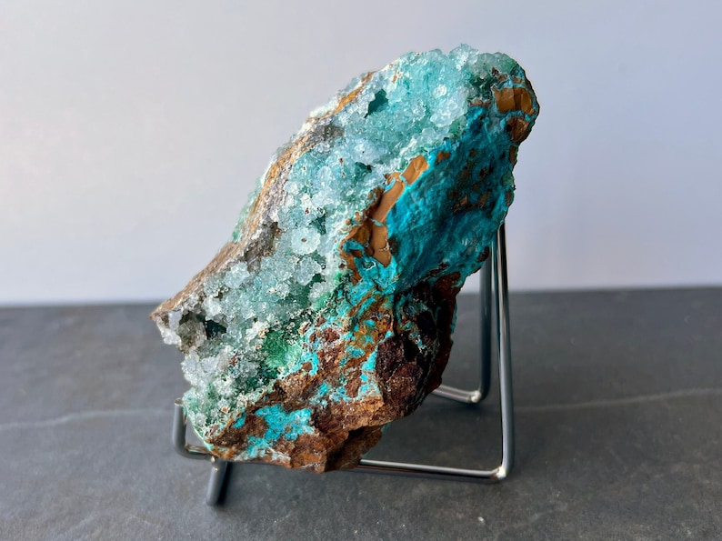 Puede incluir: Un esp&eacute;cimen de cristal azul verdoso y marr&oacute;n con formaciones de cristales blancos. El cristal est&aacute; en un soporte de metal.