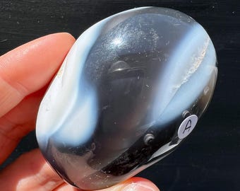 Könnte beinhalten: Ein polierter, oval geformter Stein mit einem wirbelnden Muster aus schwarzen und weißen Bändern. Der Stein ist glatt und reflektierend, mit glänzender Oberfläche. Ein kleiner weißer Aufkleber mit dem Buchstaben "A" ist angebracht.