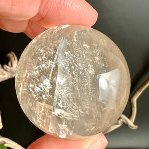 May include: A clear, spherical crystal ball with internal fractures and inclusions. The polished surface reflects light, creating a shimmering effect. The crystal is held in a hand, showcasing its size and clarity. A natural, decorative item.