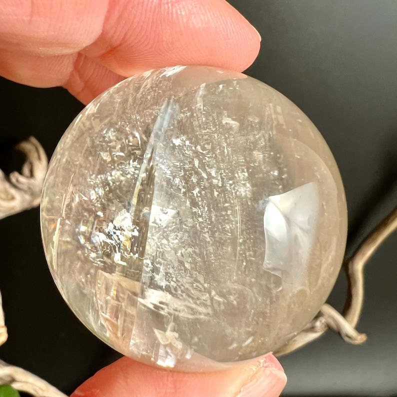 May include: A clear, spherical crystal ball with internal fractures and inclusions. The polished surface reflects light, creating a shimmering effect. The crystal is held in a hand, showcasing its size and clarity. A natural, decorative item.