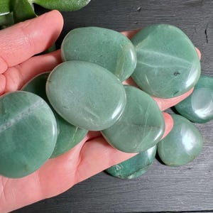 Pode incluir: Uma coleção de pedras de palma de aventurina verde, lisas e ovais. As pedras, em tons variados de verde, têm um acabamento polido e brilhante. São seguradas em uma mão, destacando sua beleza natural e textura. Cada pedra mede aproximadamente 5-7 cm de comprimento.