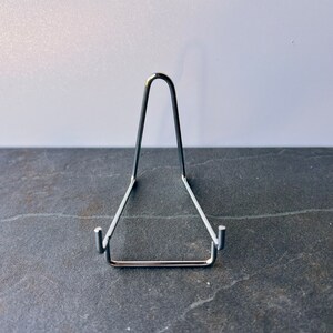 Puede incluir: Un soporte de exhibici&oacute;n de metal plateado para un plato u otro objeto. El soporte tiene forma de tri&aacute;ngulo con una parte superior curva.