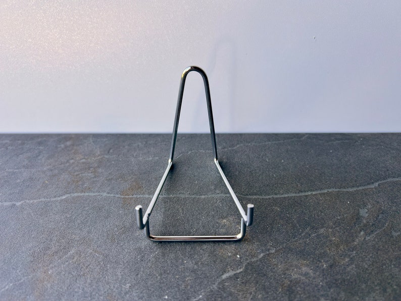Puede incluir: Un soporte de exhibici&oacute;n de metal plateado para un plato u otro objeto. El soporte tiene forma de tri&aacute;ngulo con una parte superior curva.