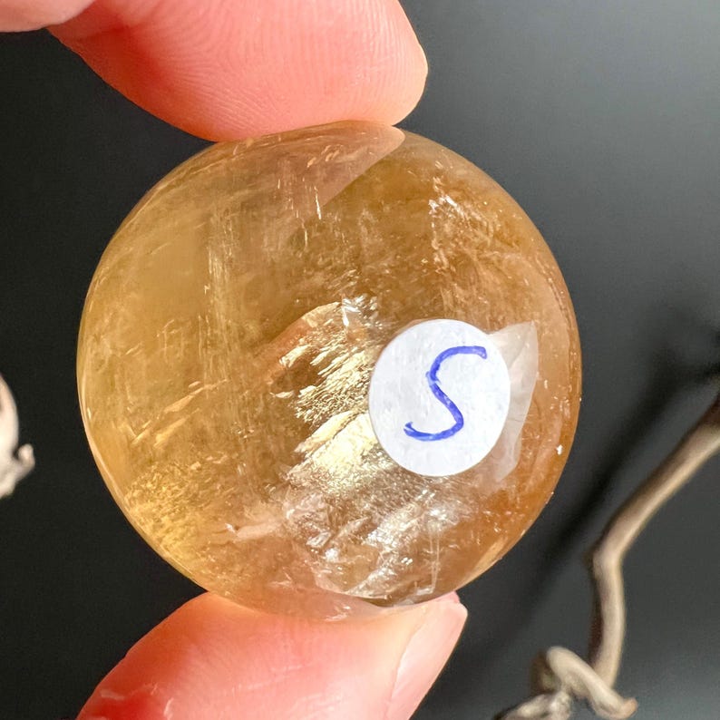 May include: A golden-yellow, translucent crystal sphere. A small white sticker with a blue "S" is affixed to the surface. The sphere is held between fingers against a dark background, with a piece of wood visible.