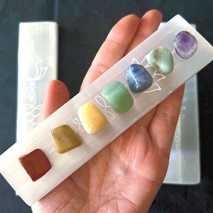 May include: A white selenite charging plate with seven chakra stones in a rainbow order. The stones are red, orange, yellow, green, blue, indigo, and violet. The plate has a lotus flower design.